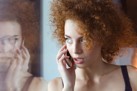 Beautiful thoughtful curly young redhead woman standing at the window and talking on cell phoneの写真素材