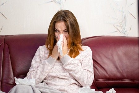 Sick ill young woman sitting on sofa in pajamas and using handkerchief for her noseの写真素材