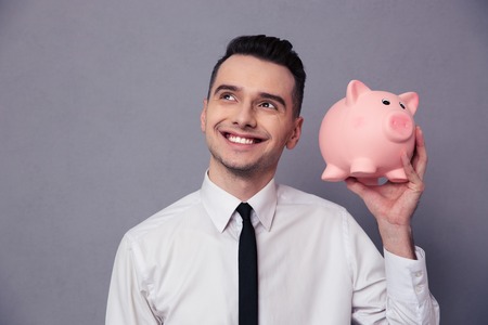 Portrait of a happy businessman holding pig money box over gray backgroundの写真素材