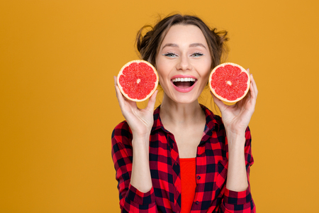 Portrait of smiling beautiful young woman in checkered shirt holding two halves of grapefruit over yellow backgroundの写真素材
