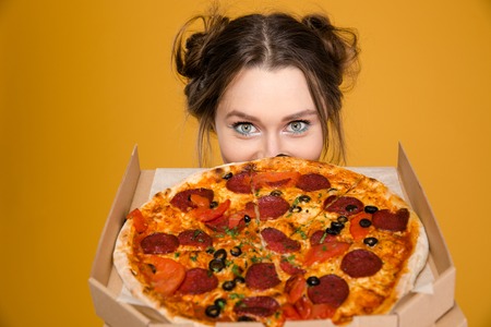 Lovely cute positive young woman hiding behind pizza over yellow backgroundの写真素材