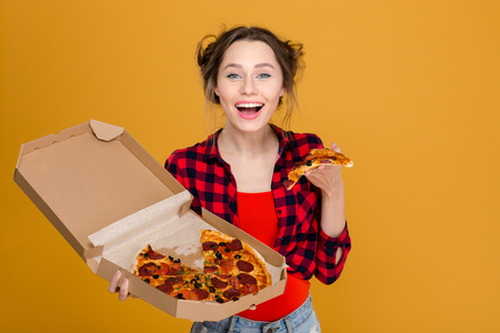 Cheerful pretty young woman in plaid shirt enjoying pizza over yellow backgroundの写真素材
