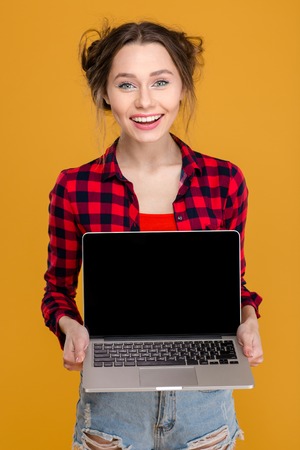 Happy attractive young woman in checkered shirt standing and holding blank screen laptop over yellow backgroundの写真素材