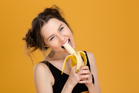 Portrait of funny beautiful young woman eating banana over yellow backgroundの写真素材