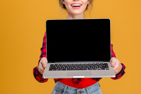 Cheerful cute lovely young woman in plaid shirt showing blank screen laptop over yellow backgroundの写真素材