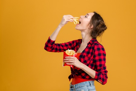 Profile of beautiful happy woman in plaid shirt tasting french fries over yellow backgroundの写真素材