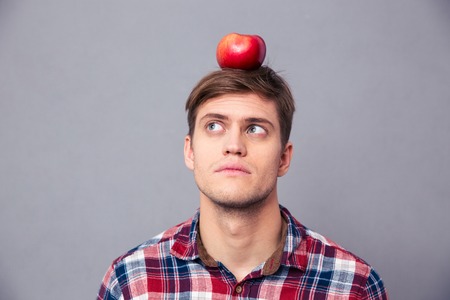 Thoughtful concentrated young man in checkered shirt with apple on his head over grey backgroundの写真素材