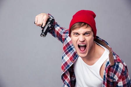 Dangerous agressive young man in checkered shirt and red hat shouting and threatening with gun over grey backgroundの写真素材