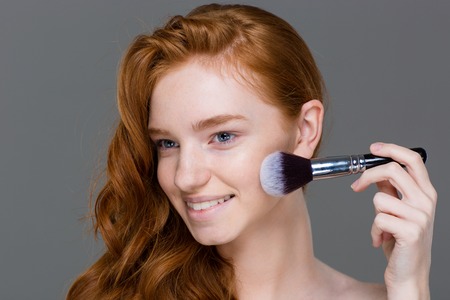 Smiling lovely young female with beautiful long hair applying powder blush brush for makeup over grey backgroundの写真素材