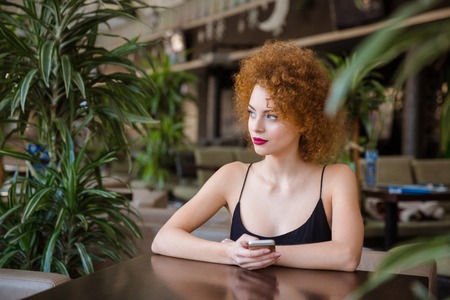 Thoughtful redhead woman with curly hair sitting at the table with smartphone in restaurant and...