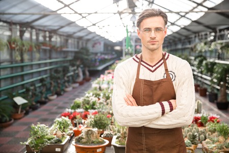 Confident man gardener in brown apron and glasses standing in greenhouse with arms crossedの写真素材