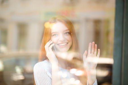 Portrait of a smiling redhead woman talking on the phone and waving palm to somebodyの写真素材