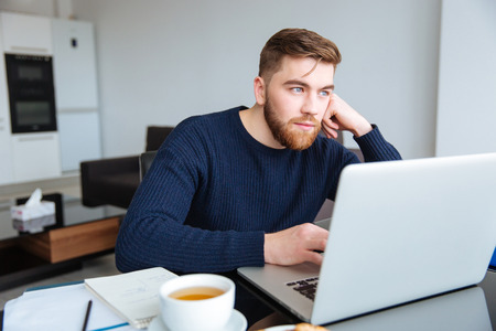 Portrait of a pensive man sitting at the table with laptop computer at homeの写真素材