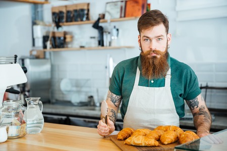 Attractive young male barista with bearrd and tattoo standing in coffe shop with croissantsの写真素材