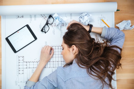 Top view of tired young woman lying and sleeping on blueprintの写真素材