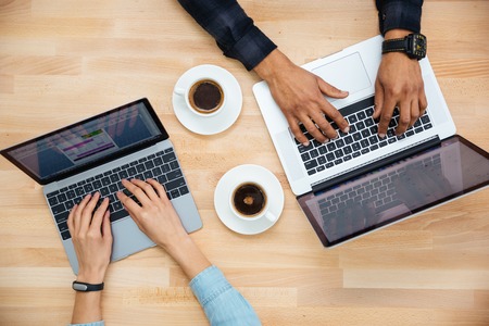 Top view of hands of african man and caucasian woman typing on two laptops and drinking coffee on wooden tableの写真素材