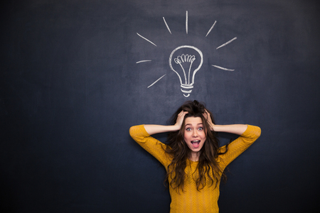 Excited pretty young woman with tousled hair and hands on head standing over blackboard background
