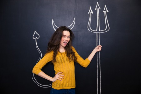 Flirty young woman standing and winking over chalkboard background with drawn devils horns and...