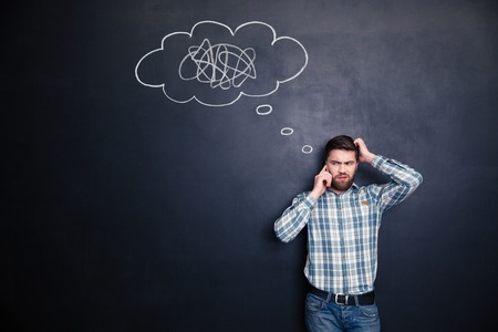 Irritated young man with beard solving a problem using mobile phone standing over blackboard...