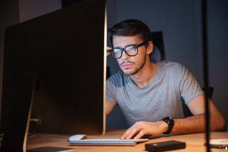 Thoughtful young man in glasses thinking and working with computer in dark room