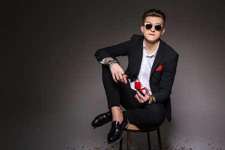 Young man in trendy cloth sitting on the chair and holding box with a proposal ring over black backgroundの写真素材