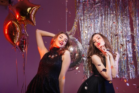 Two charming playful young women in black dresses dancing and having party over purple backgroundの写真素材