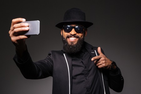 Happy afro american man in fashion cloth making selfie photo on smartphone over black backgroundの写真素材