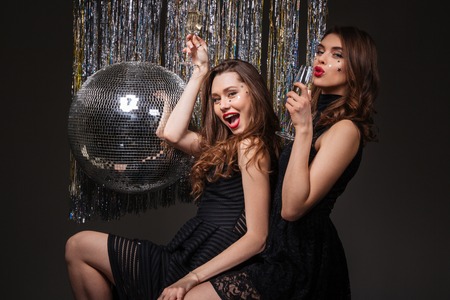 Two joyful pretty young women celebrating and drinking champagne over black backgroundの写真素材
