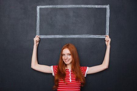 Attractive smiling redhead young woman holding a frame drawn on blackboard background above her headの写真素材