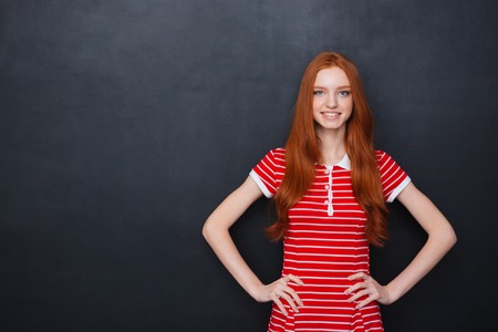 Cheerful beautiful redhead young woman smiling with hands on waist over blackboard backgroundの写真素材