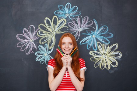 Happy beautiful young woman holding colorful pencils and standing over blackboard background with drawn flowersの写真素材