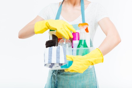 Closeup of cleaning supplies in plactic box holded by young housewife in blue kitchen apron over white backgroundの写真素材