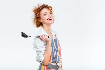 Smiling redhead housewife with curly hair in apron holding soup ladle isolated on a white backgroundの写真素材
