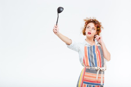 Happy redhead housewife with curly hair in apron looking on soup ladle isolated on a white backgroundの写真素材