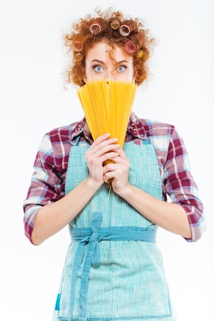 Portrait of amazed pretty young housewife with curlers in blue apron covered face with uncooked spagetti over white backgroundの写真素材