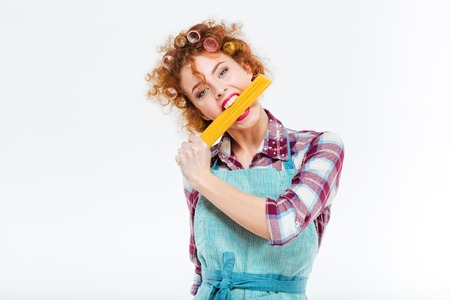 Pretty redhead housewife in kitchen apron standing and posing with uncooked spagetti over white backgroundの写真素材