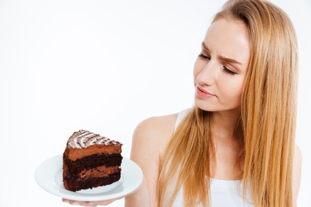 Pensive beautiful young woman looking at piece of chocolate cake over white backgroundの写真素材