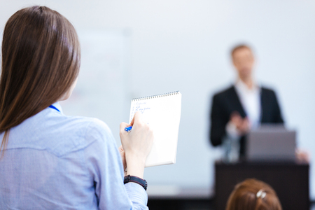 Back view of young business woman writing and making notes in notepad sitting on public presetration...