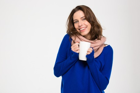 Cheerful attractive young woman in warm pink scarf standing and holding white mug with hot tea over white backgroundの写真素材