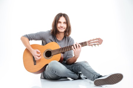Smiling young man with long hair sitting on the floor and playing guitar over white backgroundの写真素材