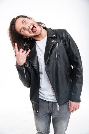 Crazy young man in black leather jacket showing tongue and doing rock gesture over white backgroundの写真素材