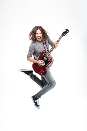 Happy young man with long hair jumping and playing electric guitar over white background