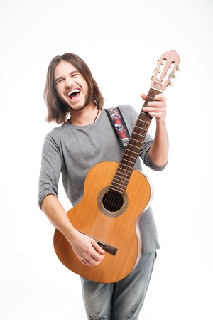 Excited handsome young man with long hair laughing and playing acoustic guitar over white backgroundの写真素材