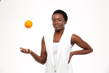 Happy playful african american young woman thowing orange up in the air over white backgroundの写真素材