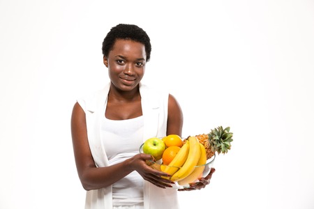 Beautiful cheerful african american young woman standing and holding glass bowl with exotic fruits over white backgroundの写真素材