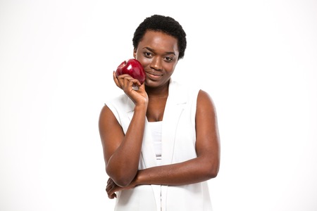 Beautiful smiling african american young woman standing and holding red apple over white backgroundの写真素材