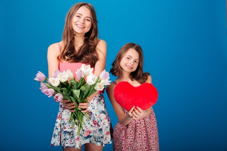Two cheerful cute sisters holding bouquet of flowers and heart over blue backgroundの写真素材