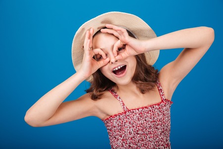 Funny playful little girl in hat making glasses by hands over blue backgroundの写真素材