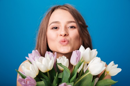 Cute lovely young woman with bouquet of tulips sending a kiss over blue backgroundの写真素材