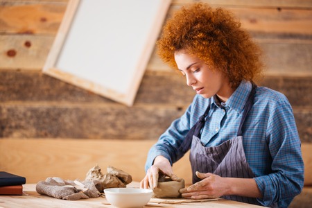 Creative young woman potter with curly red hair making earthen dishes in workshopの写真素材
